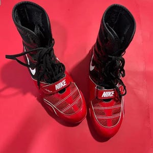 May include: A pair of high-top Nike boxing shoes in red and black. The shoes feature black laces, a white Nike swoosh logo on the side, and a red strap across the midfoot with the word NIKE printed in white. The toe area is made of red mesh.