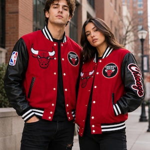 May include: Two red and black varsity jackets featuring the Chicago Bulls logo on the chest, an NBA patch on the sleeve, and the word Chicago in white script on the arm. The jackets have black leather sleeves, white snap buttons, and striped ribbed trim.