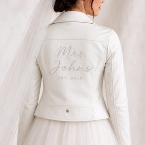 May include: A white faux leather jacket with the embroidered text "Mrs. Johns EST. 2026" in a script font. The jacket has a classic biker jacket design with a notched collar and a single gold button.