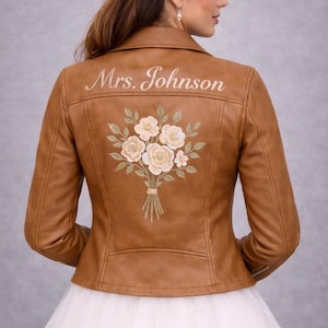 Op de afbeelding: Bruine leren jas met de geborduurde tekst "Mrs. Johnson" op de rug. Daaronder een geborduurd boeket van witte en crèmekleurige bloemen met groene bladeren. De jas heeft een klassieke biker-stijl.