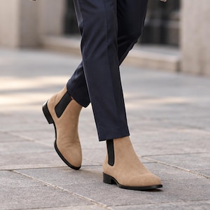 Könnte beinhalten: Ein Paar Chelsea-Boots aus beigem Wildleder mit schwarzen elastischen Seiteneinsätzen und einem flachen schwarzen Absatz. Die Stiefel werden mit einer dunkelblauen Hose kombiniert, die knapp über dem Knöchel endet und das Material zeigt.