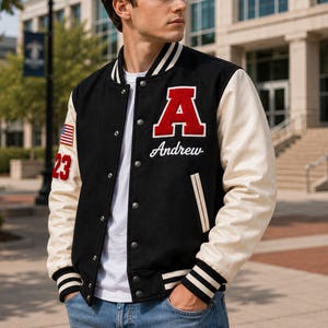 Könnte beinhalten: Eine schwarze Collegejacke mit cremefarbenen Ärmeln, verziert mit einem großen roten Buchstaben A als Aufnäher, dem in Weiß gestickten Namen Andrew, einer kleinen US-Flagge und der Zahl 23 auf dem Ärmel, kombiniert mit einem weißen T-Shirt und blauer Jeans.