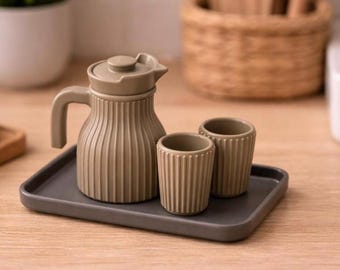 Puede incluir: Un juego de té de cerámica color topo sobre una bandeja gris. El juego incluye una tetera acanalada con tapa y asa, y dos tazas acanaladas a juego. El juego se exhibe sobre una superficie de madera clara.
