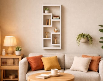 May include: A rectangular white wall-mounted shelf unit with multiple open compartments. The shelf features an asymmetrical geometric design and holds small decorative items including books, a potted plant, and gold-toned figurines.