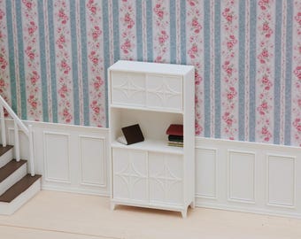 Puede incluir: Un armario de casa de muñecas blanco con dos juegos de puertas correderas con un patrón geométrico de estrella. El armario tiene un estante central abierto que contiene un pequeño libro oscuro y una pila de tres libros en miniatura.