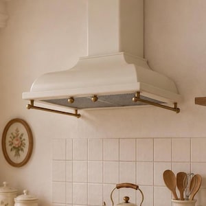 May include: A cream-colored kitchen range hood featuring a wide, flared base with a metallic rail and three decorative knobs. The hood has a central chimney section and a mesh filter underside, mounted above a tiled wall in a kitchen setting.