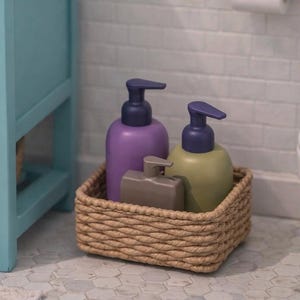 May include: A wicker basket contains three soap dispensers in a bathroom. Two are rounded, one purple and one green, with dark blue pump tops. A smaller, rectangular dispenser is also present. The basket sits near a blue cabinet and a white toilet.