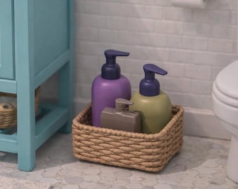 Puede incluir: Una cesta cuadrada de cuerda trenzada que contiene tres botellas de plástico con dosificador. Las botellas son de color morado, verde oliva y gris, cada una con un mecanismo de bomba azul oscuro o gris. La cesta tiene un aspecto trenzado.
