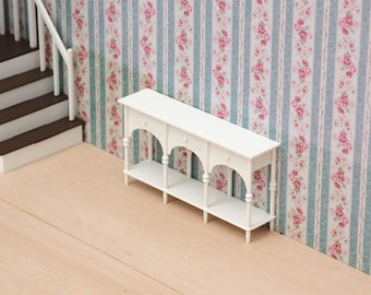 Puede incluir: Una mesa consola en miniatura de color blanco que presenta tres cajones pequeños con pomos redondos, recortes arqueados y un estante inferior. La pieza está construida con patas delgadas y torneadas, situada contra una pared con papel pintado floral.