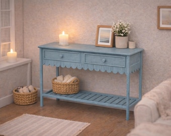 Puede incluir: Una mesa consola de madera azul claro con dos cajones con pomos redondos, un faldón festoneado y un estante inferior de listones. La mesa rectangular se apoya sobre cuatro patas rectas y muestra un acabado desgastado con veta de madera.