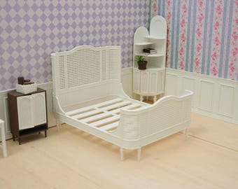 Puede incluir: Un marco de cama en miniatura blanco con cabecero y piecero de rejilla tejida, acompañado de un pequeño armario marrón con puertas blancas, una silla blanca y una estantería de esquina blanca con una pequeña planta en maceta.