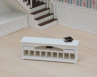 Puede incluir: Una consola multimedia en miniatura de madera blanca con dos cajones pequeños, un estante central abierto y ocho compartimentos de almacenamiento verticales. Tres libros en miniatura de color oscuro están sobre la consola y el estante.