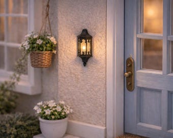 Puede incluir: Un farol de pared en miniatura de color negro con una luz encendida, situado entre una puerta azul claro con pomo de latón y una ventana. Una cesta trenzada colgante y una maceta blanca con pequeñas flores blancas completan la escena.