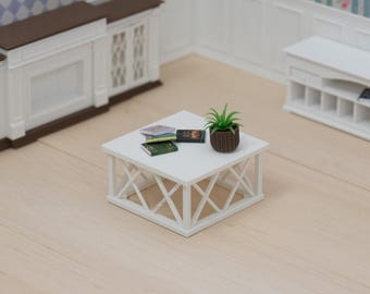 Puede incluir: Una mesa de centro cuadrada en miniatura de color blanco con una base de patrón cruzado. Sobre la mesa hay una pequeña maceta marrón estriada con una planta verde y una pila de tres libros diminutos con portadas coloridas en tonos verde y morado.