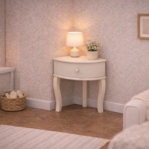 May include: A cozy corner scene featuring a small, off-white nightstand with a drawer, a lamp, and a potted plant. A lit candle and a window are visible. A woven basket and a framed picture complete the warm, inviting decor.