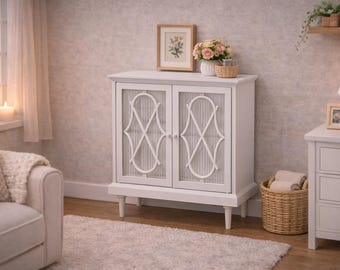 Puede incluir: Un armario de almacenamiento blanco con dos puertas que presentan paneles de vidrio estriado vertical y un diseño decorativo blanco superpuesto con un patrón geométrico curvo y simétrico. El mueble se apoya sobre cuatro patas cónicas y tiene dos pomos redondos blancos.