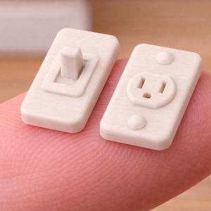 May include: Miniature off-white electrical outlet and light switch. The outlet has two slots and a grounding hole, with two small circular details. The light switch has a rectangular toggle. Both are small, detailed replicas.