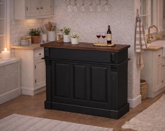 Puede incluir: Una isla de cocina negra con una encimera de madera oscura. La isla tiene un frente panelado y está decorada con una botella de vino, dos copas de vino y una pequeña bandeja. La cocina tiene armarios blancos y una ventana.