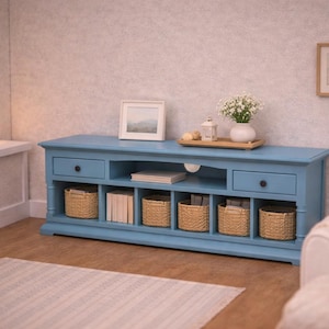 Könnte beinhalten: Hellblaues TV-Möbel mit zwei Schubladen und offenen Staufächern. Geflochtene Körbe und Bücher werden in den Fächern aufbewahrt. Ein gerahmtes Bild und eine Vase mit Blumen stehen oben drauf. Ein Teppich und ein weißer Stuhl befinden sich im Raum.