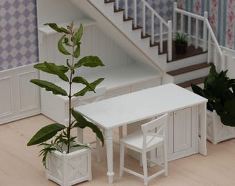 Puede incluir: Un conjunto de comedor en miniatura para casa de muñecas que incluye una mesa rectangular blanca con armario integrado, dos sillas blancas con respaldo cruzado y dos maceteros cuadrados blancos con plantas verdes junto a una escalera blanca.
