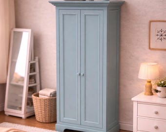 May include: A tall, light blue wooden wardrobe featuring two vertical doors with simple round knobs. The cabinet has a flat top with decorative molding, a recessed panel design on each door, and a sturdy base with a cutout detail at the bottom.