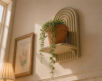 Puede incluir: Una estantería de pared arqueada de color crema con una maceta de terracota que contiene una planta verde colgante. La estantería está montada en una pared beige, con una impresión botánica enmarcada y una lámpara en el fondo. La estantería tiene un diseño decorativo.