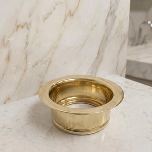 May include: A shiny, gold-colored sink drain with a wide, flat rim and a cylindrical body. The drain is made of metal and has a smooth, reflective surface. The background features a marble countertop and wall.