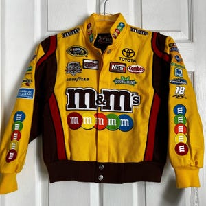 May include: A yellow racing jacket with brown and red accents. The jacket features a large M&M's logo with the text "m&m's" and a row of colorful M&M's candies. The jacket also has various sponsor logos, including Toyota, NOS, Combos, and Siemens.
