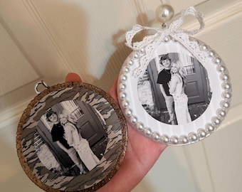 May include: Two circular ornaments featuring a black and white photograph of a couple. One ornament has a camouflage border, while the other is decorated with a white lace bow and a border of white pearls, topped with a single large pearl bead.