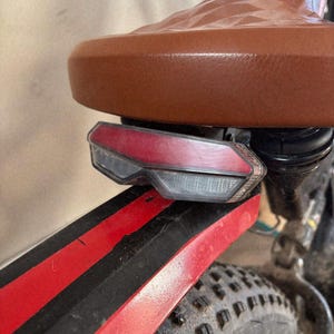 May include: Close-up of a brown leather bicycle seat with a diamond pattern. A red and clear taillight is mounted below the seat. The black fender has red stripes and a black tire is visible.