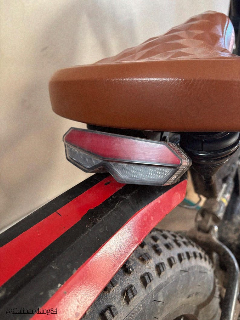 May include: Close-up of a brown leather bicycle seat with a diamond pattern. A red and clear taillight is mounted below the seat. The black fender has red stripes and a black tire is visible.