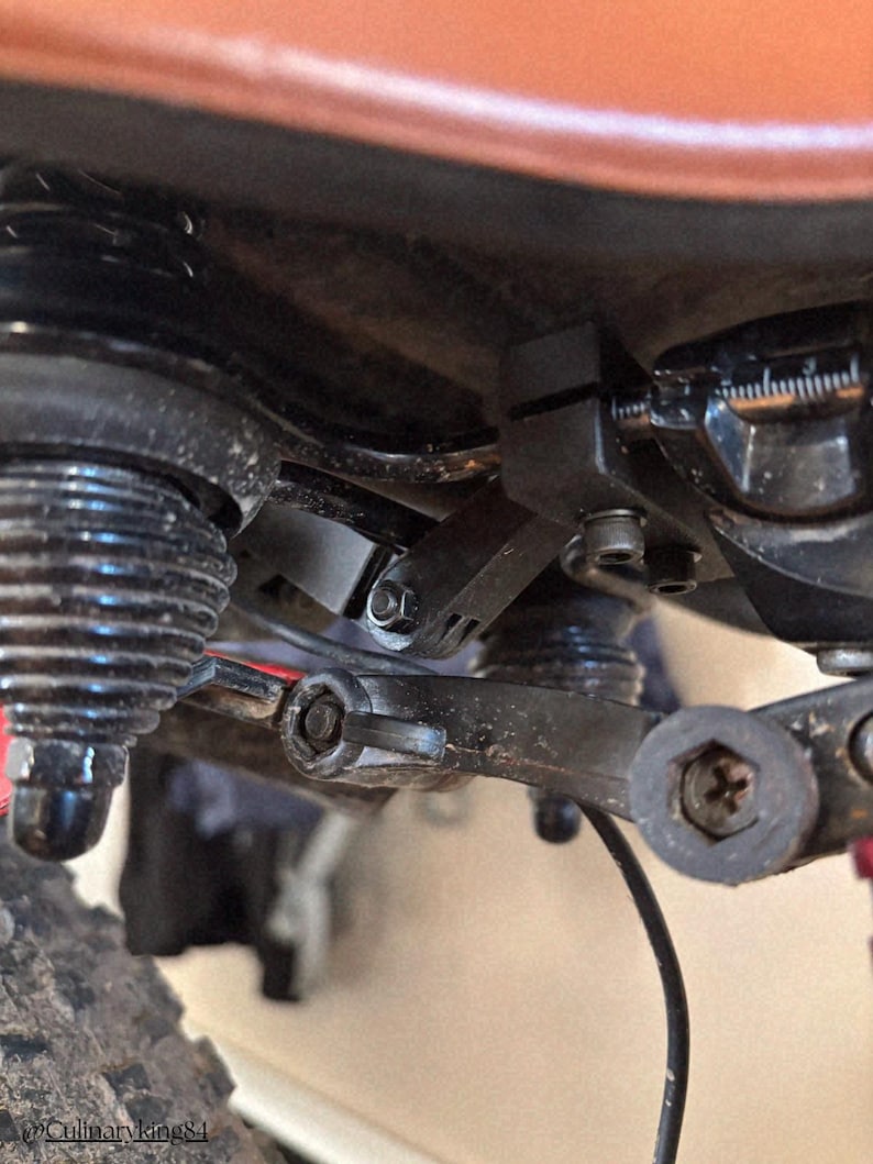 May include: Close-up of a bicycle's suspension system, showcasing black components, including a shock absorber, linkages, and bolts. The image highlights the mechanical details of the bike's frame and suspension.
