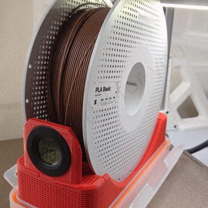 Könnte beinhalten: Eine braune PLA Basic Filamentrolle wird in einem roten und transparenten Kunststoffbehälter gehalten. Die Spule ist weiß mit Perforationen und dem Text "PLA Basic". Ein Digitaldisplay zeigt die Temperatur an.