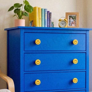 Può includere: Una cassettiera in legno blu brillante con tre cassetti orizzontali, ciascuno dotato di due pomelli rotondi gialli decorati con icone di faccine sorridenti nere. La superficie ospita una pianta verde in vaso, libri, una sveglia gialla e una cornice.