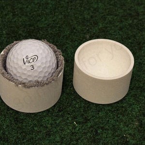 Könnte beinhalten: Ein weißer Golfball mit dem Wort "Vice" und der Zahl "3" ist in einem beigefarbenen zylindrischen Behälter mit grauem Rand platziert. Daneben befindet sich ein identischer, leerer Behälter.