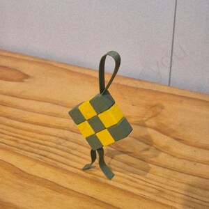 May include: A handcrafted, geometric ornament with a woven pattern of yellow and dark green squares. It has a loop for hanging and two dangling strips. The ornament is displayed on a wooden surface.