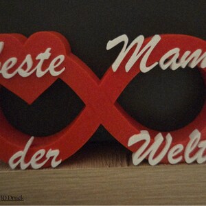 Op de afbeelding: Een rood oneindigheidssymbool met de woorden "beste Mama der Welt" in witte letters. Het ontwerp bevat een hartvorm aan de linkerkant. Het item is waarschijnlijk een decoratief stuk of een cadeau.
