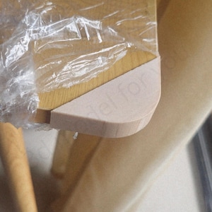 May include: Close-up of a light-colored wooden table corner, partially wrapped in clear plastic. The table has a rounded edge and a light beige finish. The table leg is visible in the background.