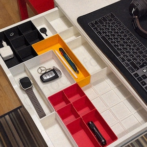 May include: An open drawer with various compartments, neatly organized with tech accessories. Includes a smartwatch, car keys, pens, a charger, and a flashlight. A keyboard and headphones are on the adjacent surface. The drawer is white with black, yellow, and red dividers.