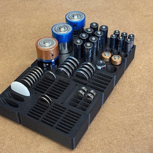 Op de afbeelding: Een zwarte plastic batterij-organizer gevuld met verschillende batterijformaten, waaronder AA, AAA, C, D en knoopcelbatterijen. De organizer heeft meerdere compartimenten om verschillende batterijtypen in op te bergen, waardoor ze netjes worden gerangschikt. De batterijen zijn in verschillende kleuren.