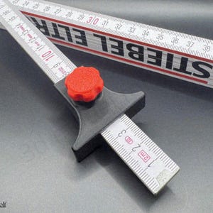 Può includere: Uno strumento di misurazione nero e rosso con un pomello rosso, utilizzato con un metro a stecca. Il metro è bianco con segni neri e la scritta "STIEBEL ELTR". Lo strumento è progettato per misurare in metri, con segni visibili fino a 3 metri.