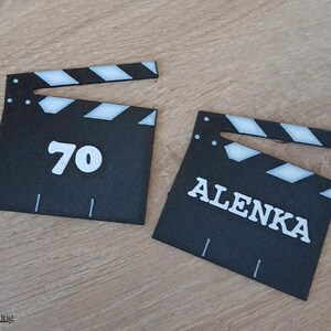 May include: Two black clapperboards with white accents and handles. One clapperboard has the number "70" in white, and the other has the name "ALENKA" in white. The clapperboards are on a wooden surface.
