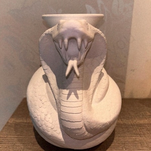 May include: A white cobra-shaped decorative vase. The vase is sculpted to resemble a cobra with its hood flared and mouth open, revealing fangs. The vase has a round base and a small, circular opening at the top. The surface has a textured appearance.