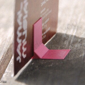 May include: A business card is propped up by a pink paper stand on a wooden surface. The card is a dark color with white text. The stand is a folded piece of paper, providing support. The image is well-lit, with shadows.