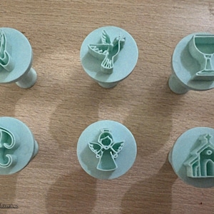 May include: Six light blue cookie stamps with religious symbols. The stamps include praying hands, a dove, a chalice, the letters "FG", an angel, and a church. Each stamp has a round base and a small handle.