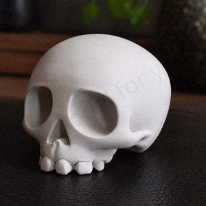 May include: A small, white skull figurine with large eye sockets and a detailed jawline. The skull is made of a smooth material and sits on a dark surface. The design is a decorative item, possibly for Halloween or gothic decor.
