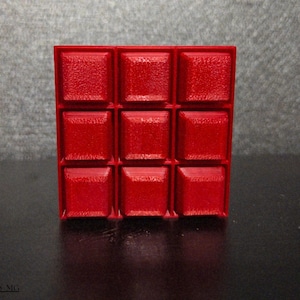 May include: A bright red, three-dimensional cube with a textured surface. The cube is made up of nine individual squares, each with a raised, square design. The overall look is modern and geometric.