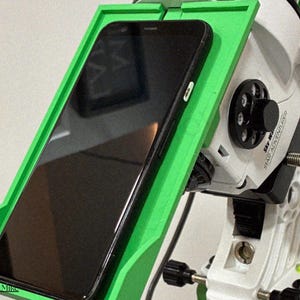 Könnte beinhalten: Eine leuchtend grüne Smartphone-Halterung, die an einer weißen Sky-Watcher Star Adventurer Teleskop-Montierung befestigt ist. Die Halterung verfügt über einen rechteckigen Rahmen, der ein schwarzes Smartphone für Astrofotografie fixiert.
