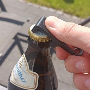 May include: A black, L-shaped bottle opener made of a textured material is positioned on the gold-colored metal cap of a brown glass beer bottle. The bottle features a white label with the text Brauhaus Reuther Hell and a wheat graphic.