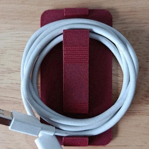 May include: A dark red rectangular cable organizer with a central strap holding a coiled white USB charging cable. The organizer features a textured surface and a secure clip design to keep the cable neatly wound and organized for storage.